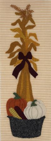 Wool applique on brushed flannel background of corn husks with a wash bucket filled with pumpkins and ears of corn at it's base.