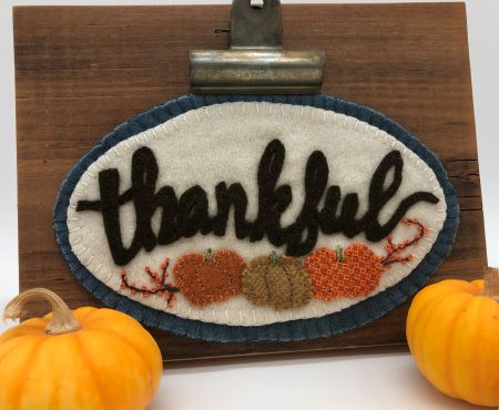 Small oval wool applique mat with the word thankful in cursive and three pumpkins underneath with a bittersweet vine.