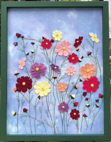 A framed rectangular wool applique wall hanging of Cosmos flower garden against a mottled blue sky. Stems and leaves are hand embroidered.