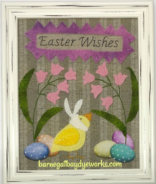 Wool applique on a pre-printed background of a chick in a bunny ears hat with pink flowers, green leaves, 5 colorful printed Easter eggs and a banner that says Easter Wishes surrounded by a wool applique purple frame. 