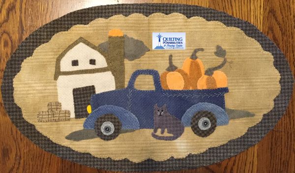 Wool applique farm scene with a barn, silo, hay bales and a vintage, blue truck filled with pumpkins and a cat sitting outside. 