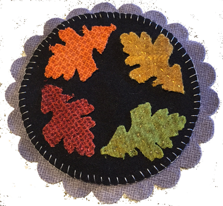 Four die cut  wool applique fall leaves in red, orange, gold and green on a black circle wool mat which is on a slightly larger gay/blue scalloped pre cut wool mat