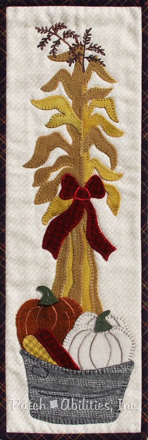 A rectangular wool applique wall hanging pattern with corn stalks, pumpkins and corn in a gray basket.