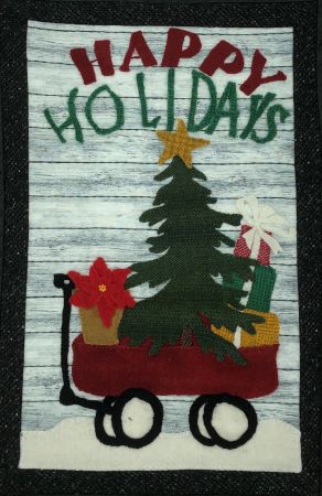Wool applique on cotton scene of a vintage red wagon with a Christmas tree, gifts and a poinsettia in it. Happy Holidays spelled out above.