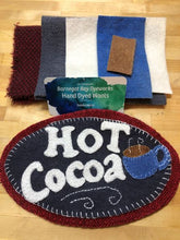 Load image into Gallery viewer, Wool Applique oval mat with &quot;Hot Cocoa&quot; in white wool and a mug in blue wool on a black background set on a larger red mat.
