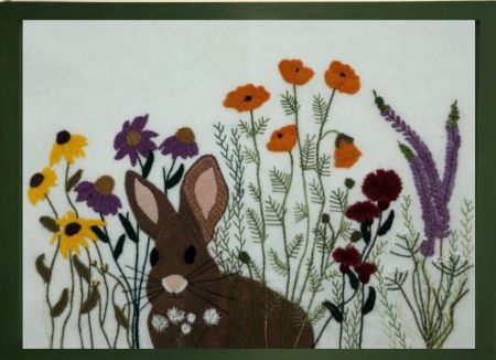 Wool applique scene of a brown rabbit in a field of flowers eating clover.  This pieces uses hand dyed wool for applique, hand embroidery and a hand dyed cotton background.