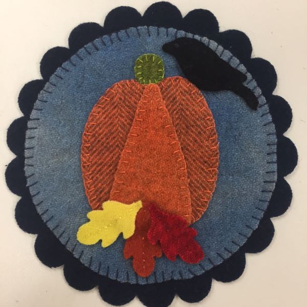 Wool Applique mat with pumpkin, leaves and a crow on a blue circle background on a slightly larger navy scalloped mat.