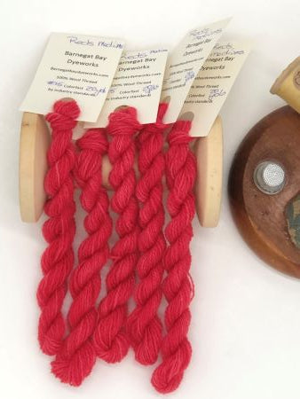 Five skeins of hand dyed, variegated, medium reds wool threads draped over a vintage thread spool.  Size 15 thread, similar to thickness of #12 pearl cotton.
