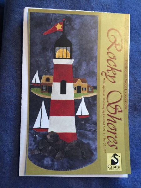 Rocky Shores wool applique scene with a prominent, red and white lighthouse protecting boats from the rocks with an island in the background.