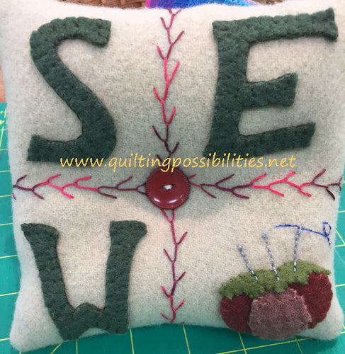 SEW Wool Pincushion Kit with feather stitch separating the square pincushion into 4 sections with SEW spelled out in green wool applique and a wool applique pincushion in the bottom right hand corner