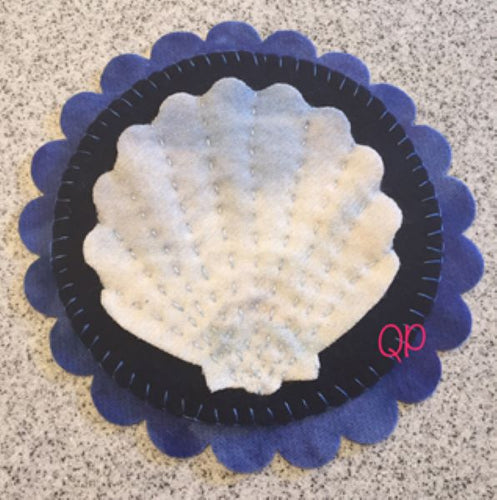 Wool applique mat with a tan/bluish scallop shell on a blue round mat on a slightly larger scalloped navy mat.