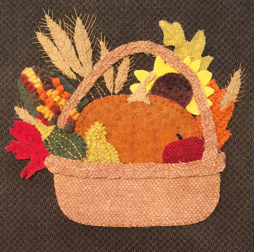 Wool applique basket of plenty  has a tan basket with handle with a pumpkin, sunflower head, fall leaves, wheat stalks, 3D Indian corn, gourds and an apple all in Fall colors.